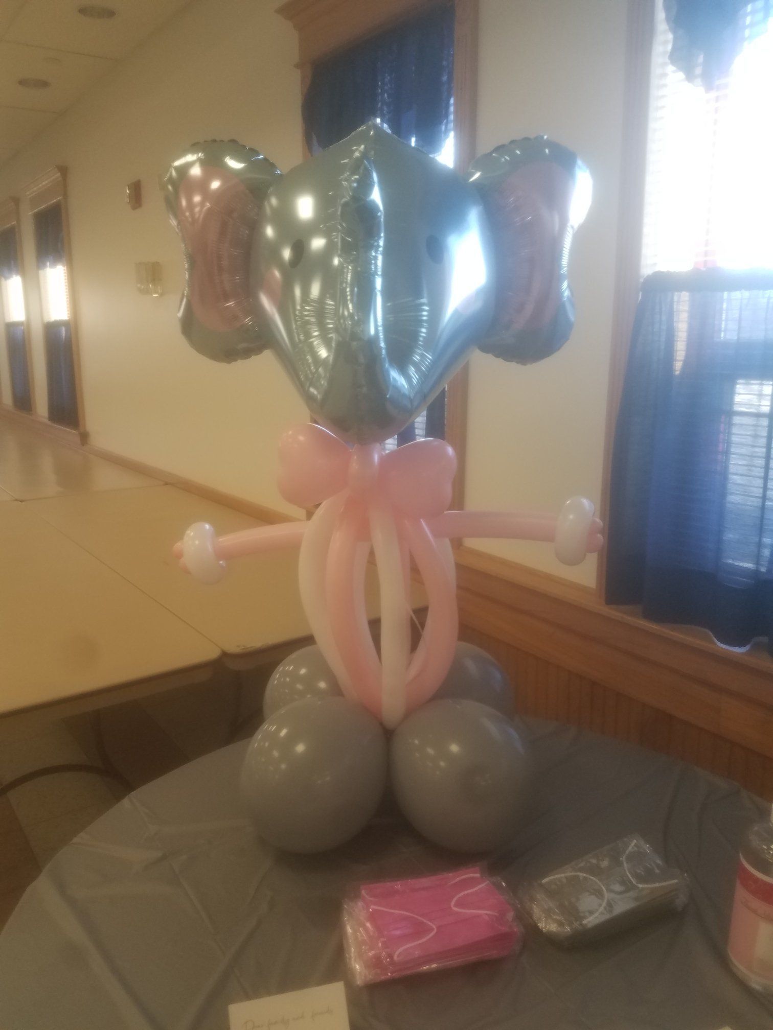Elephant-shaped balloon sculpture with pink accents on a table in a hallway, with gift boxes nearby