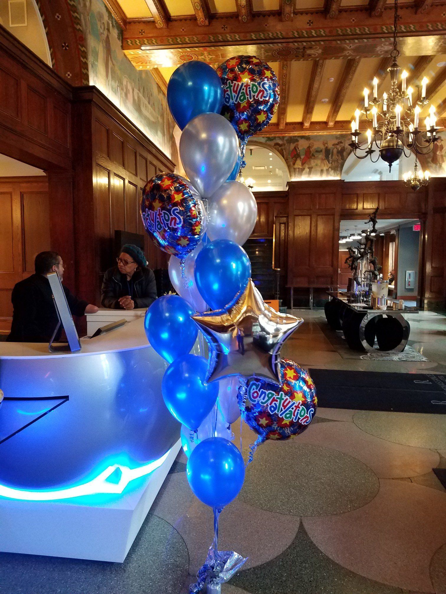 Blue and silver balloon arrangement in a wood-paneled hall with chandeliers and a glowing table nearby