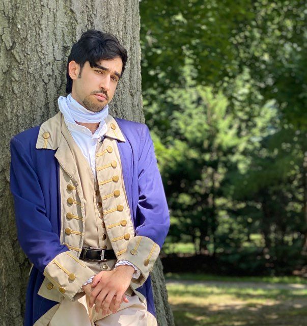 Man in a blue and cream historical-style suit leaning against a tree in a park