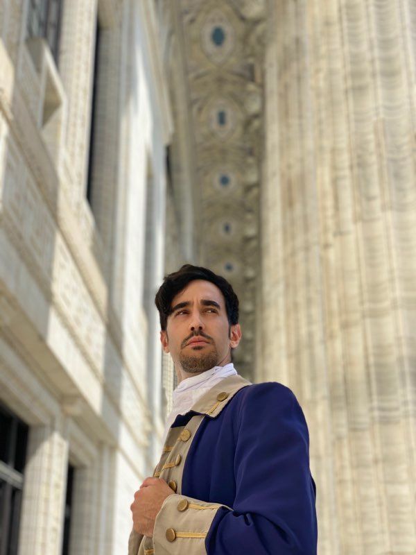 Man in a blue coat standing beneath ornate stone columns, looking upward.