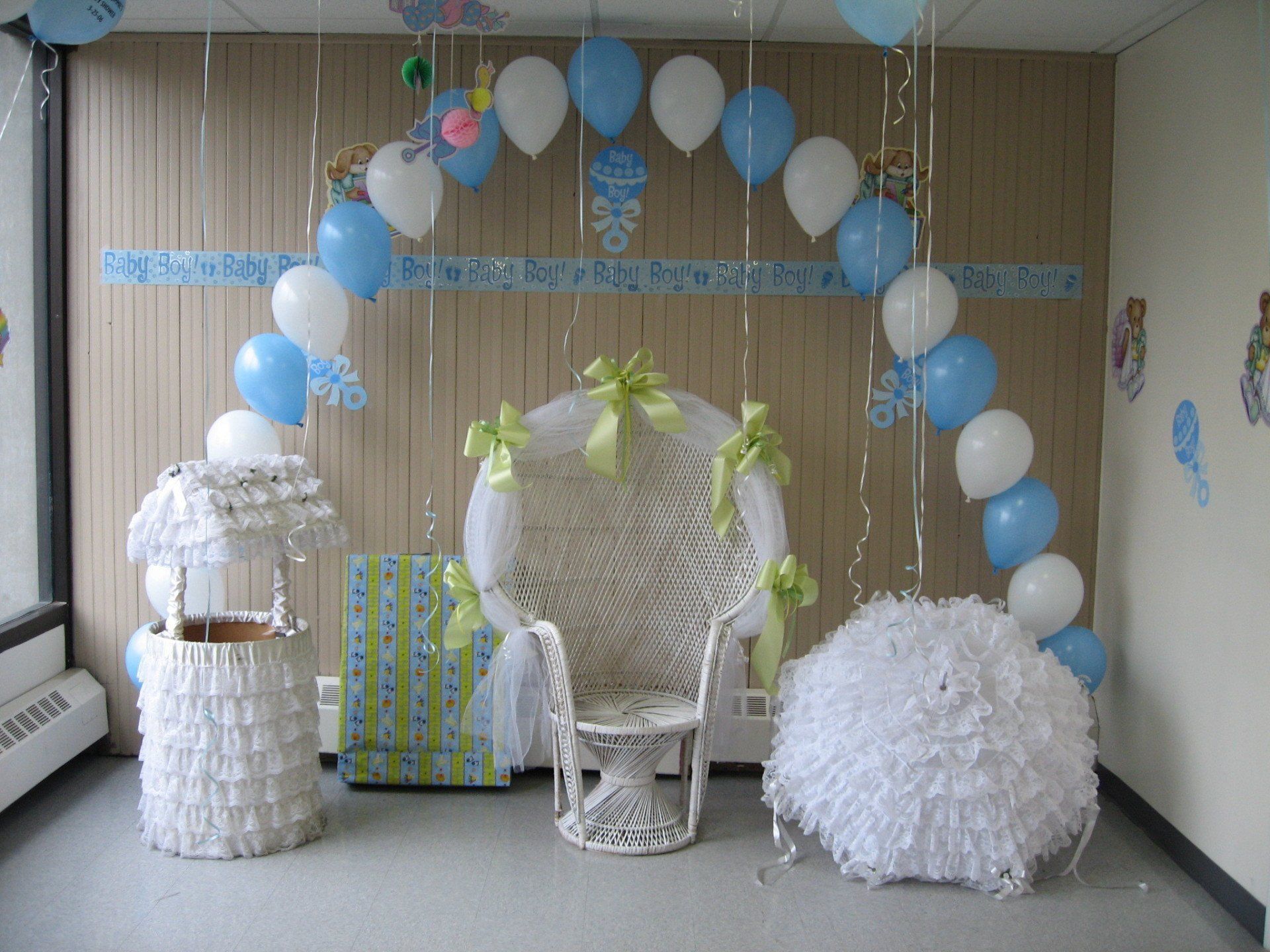 White and blue balloon arch over a decorated dessert table with a wicker chair and gifts against wood-paneled walls