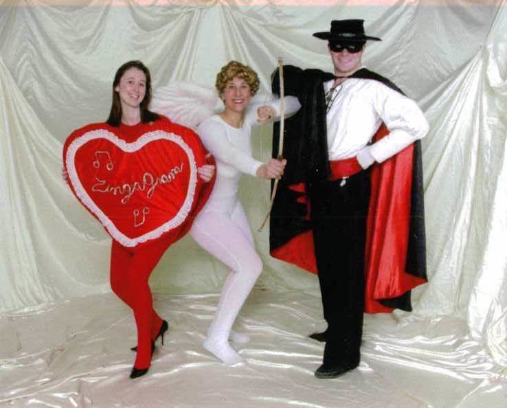 Three people posing in costumes against a white draped backdrop, including a red heart and a black cape.
