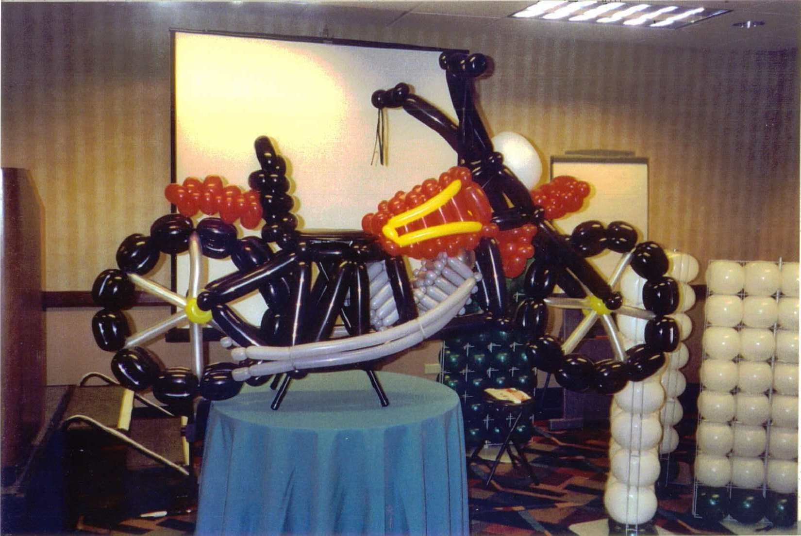 Balloon bicycle sculpture displayed indoors on a table, with black, orange, and white balloons.