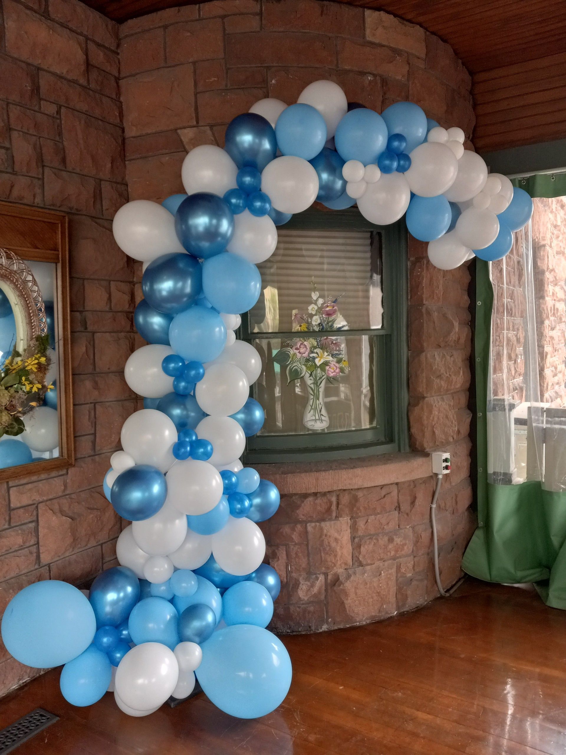 Blue, white, and silver balloon arch framing a window in a wood-paneled interior