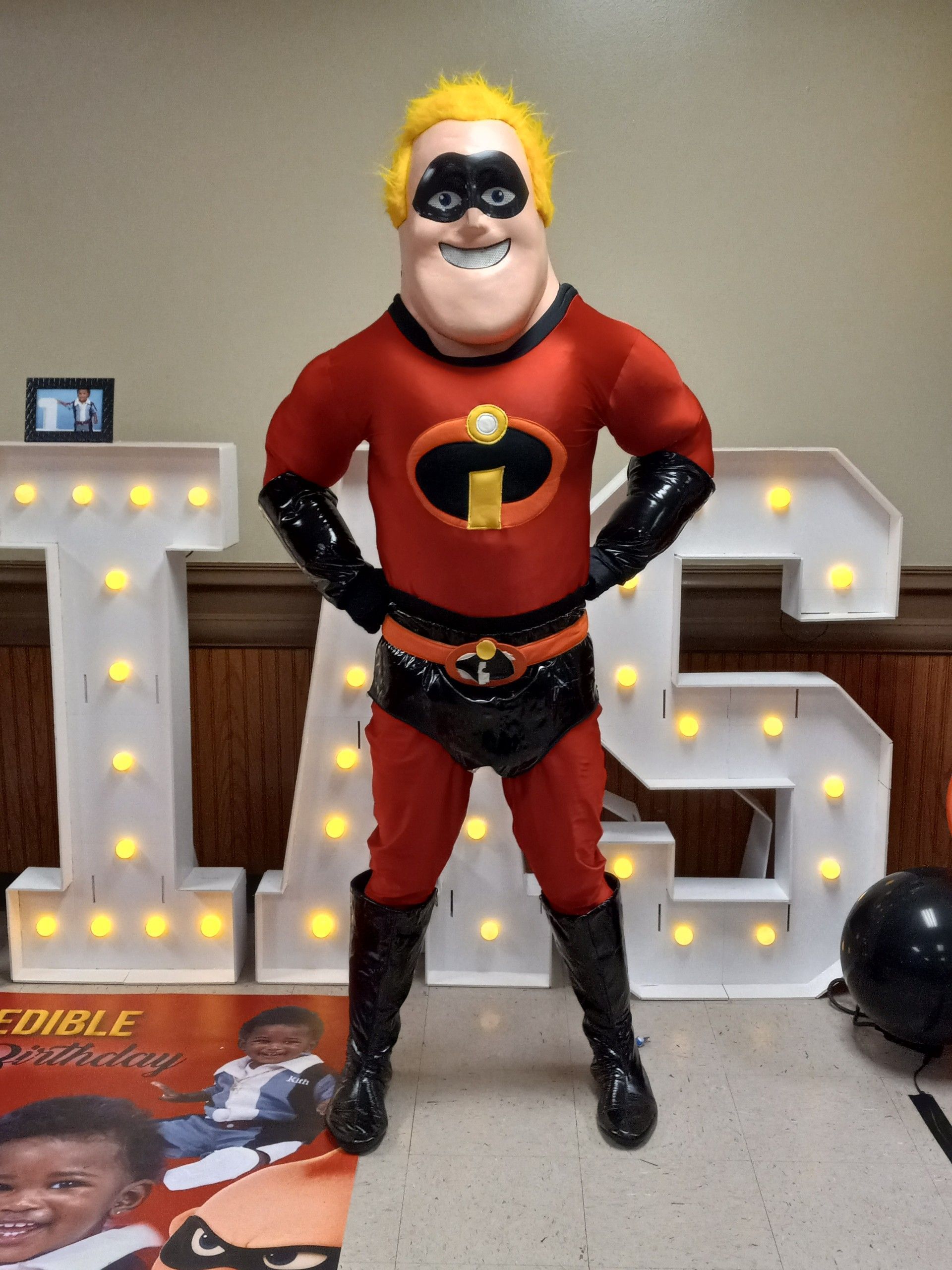 Person dressed as Mr. Incredible stands in front of lit letters with a framed photo and balloon.