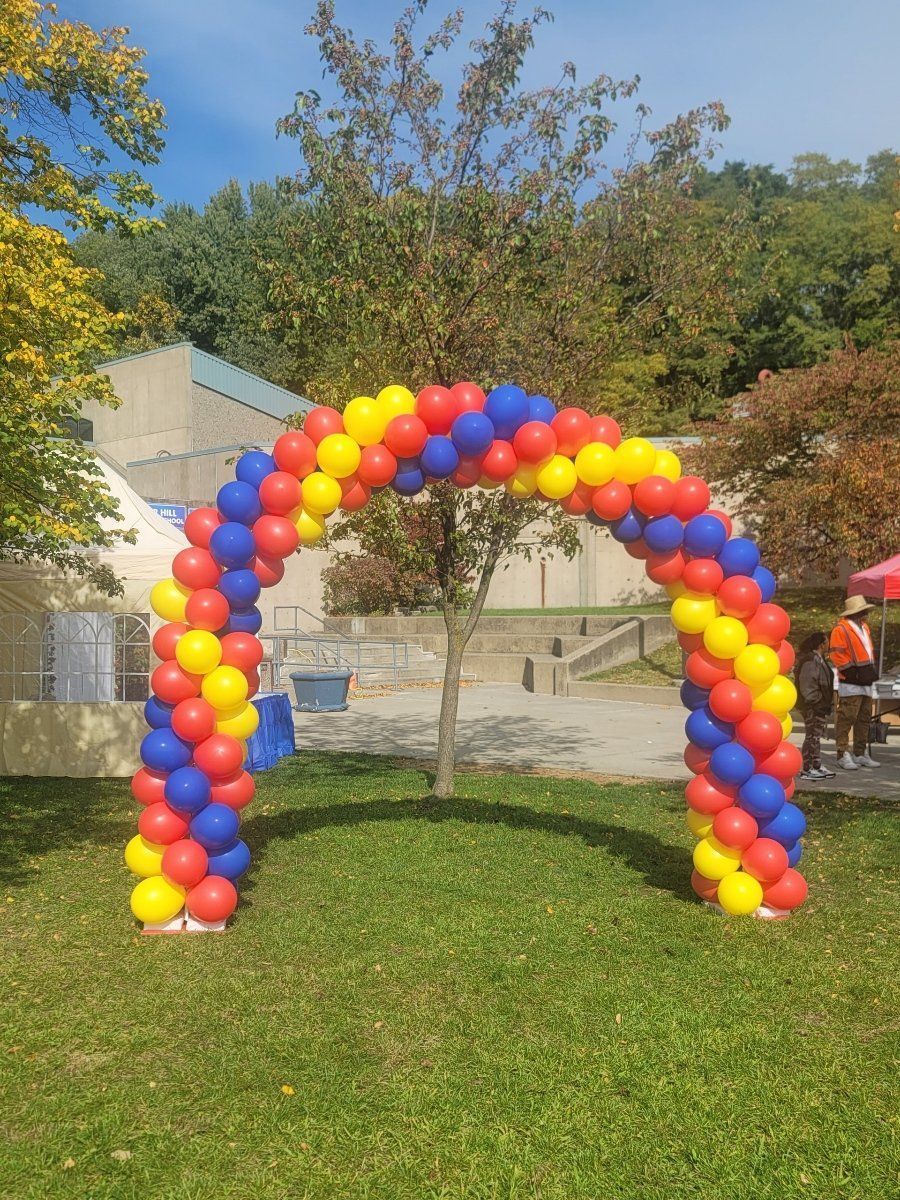 Rainbow balloon arch on a grassy lawn outdoors with trees and buildings in the background