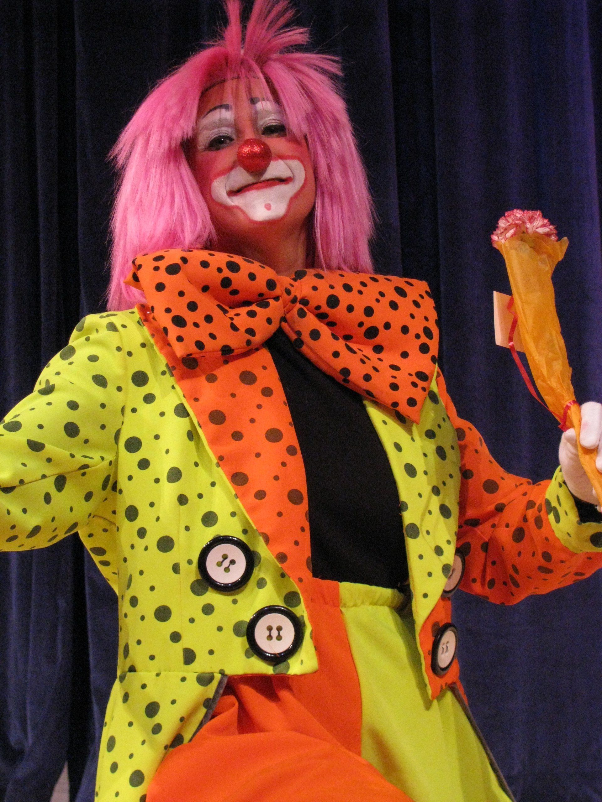 Clown in bright polka-dot costume with pink wig and oversized bow, smiling onstage against dark curtains