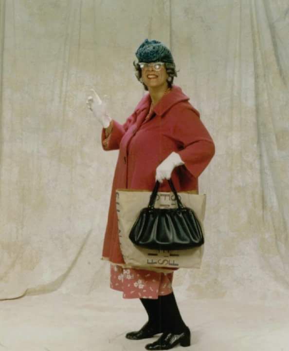 Person posing in a red coat and black hat, holding a handbag against a beige backdrop.