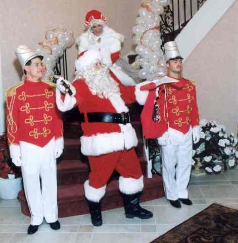 Santa in red suit posing with two white-clad performers indoors at a holiday display