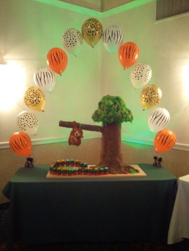 Decorated table display with a teddy bear, tree, and arch of colorful hanging balloons in a lit room