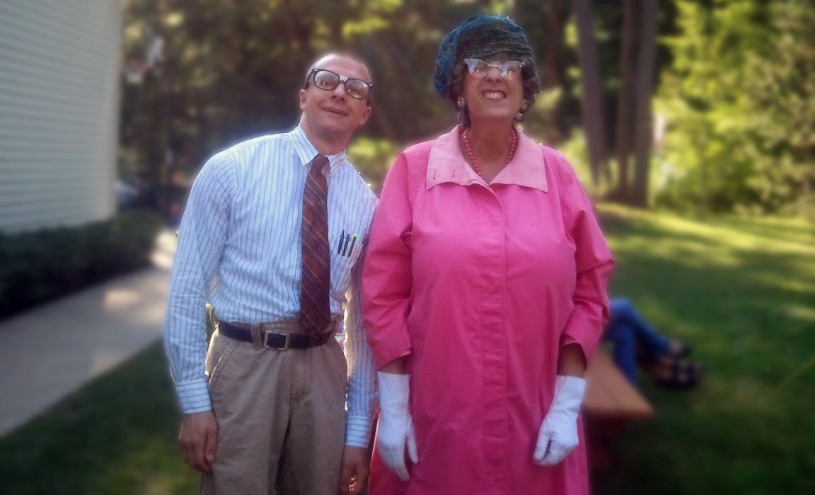 Two people standing outdoors, smiling, one in a blue striped shirt and tie, the other in a bright pink dress.