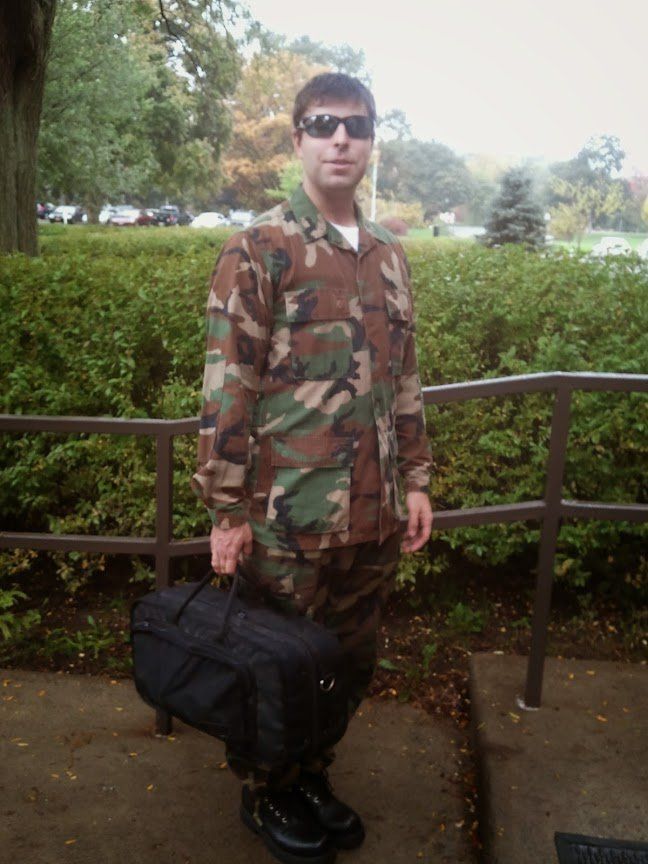 Person in camouflage clothing and sunglasses stands outdoors holding a black duffel bag near a railing.