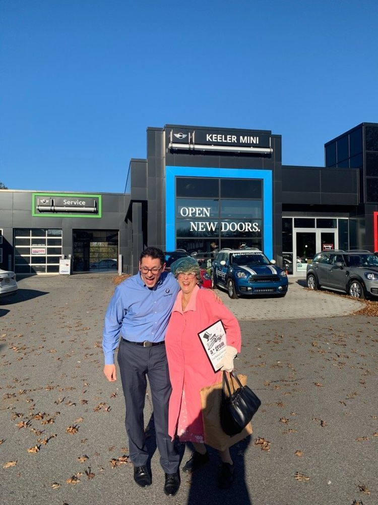 Two people posing outside a car dealership with a blue car and an open showroom sign.