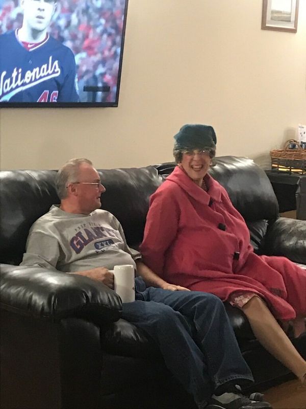 Two people sit on a black leather couch in a living room, wearing casual clothes and smiling at each other.