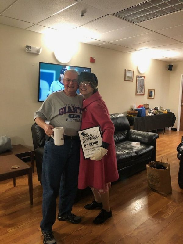 Two people smiling and hugging in a living room, one holding a mug and a sign.