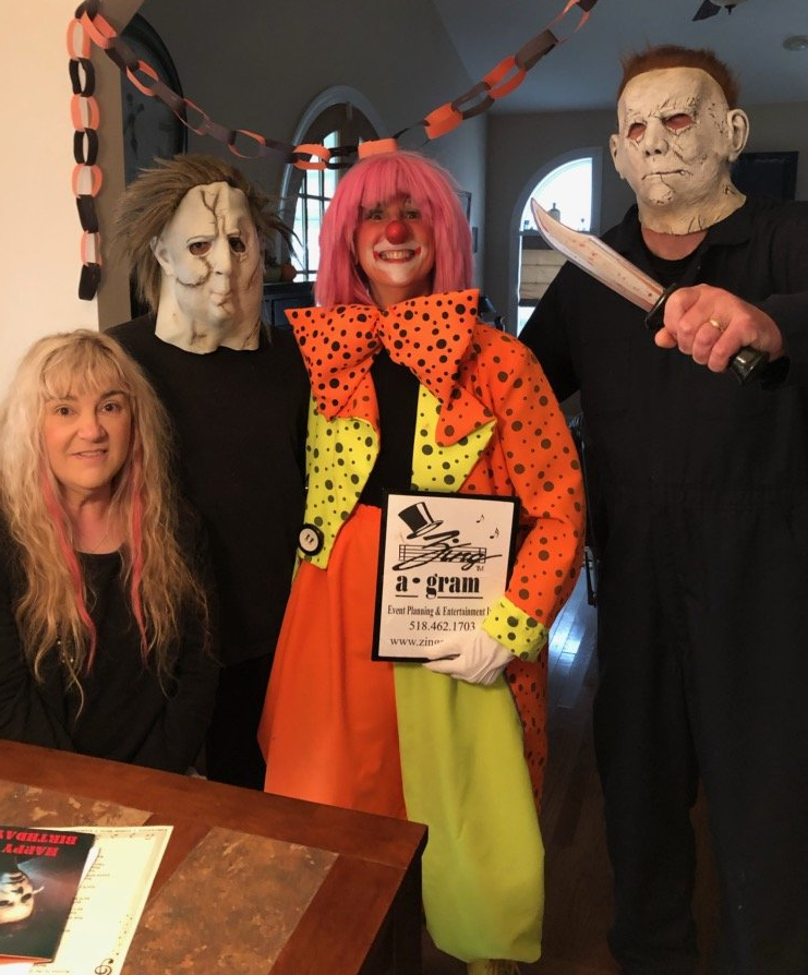 Four people in Halloween costumes pose indoors, including masked figures and a clown holding a sign.