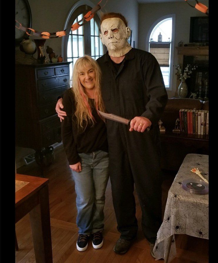 Person and Michael Myers costume pose together in a dim living room, holding a knife.