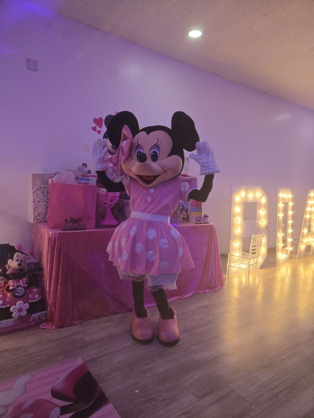 Minnie Mouse mascot posing in a pink dress beside glowing marquee letters in a decorated room