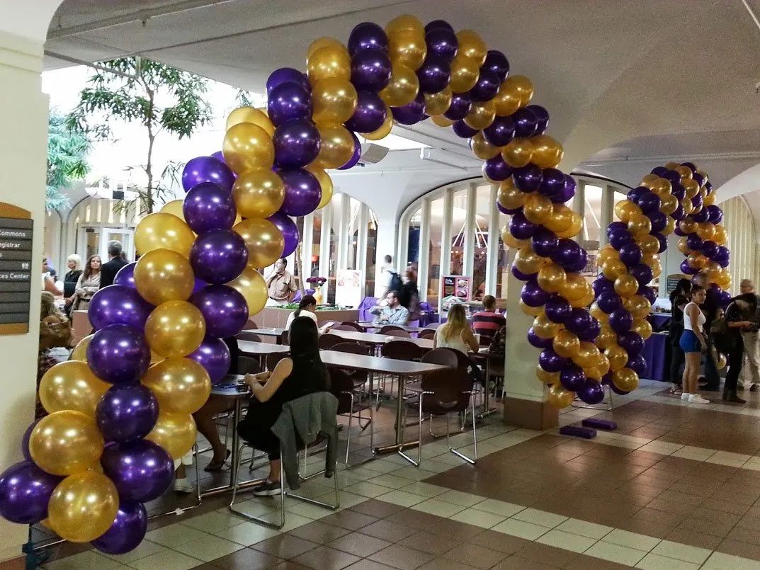 Purple and gold balloon archway in an indoor event hall with tables and seated guests