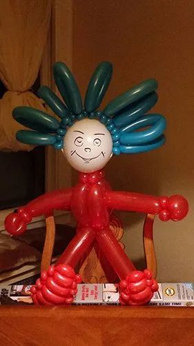 Red balloon figure with blue spiky hair and a smiling face, standing indoors on a table.
