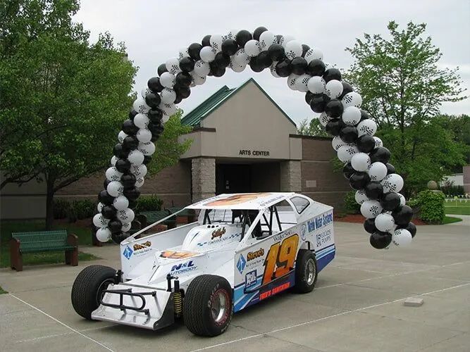 White and blue race car under black-and-white balloon arch outside a building