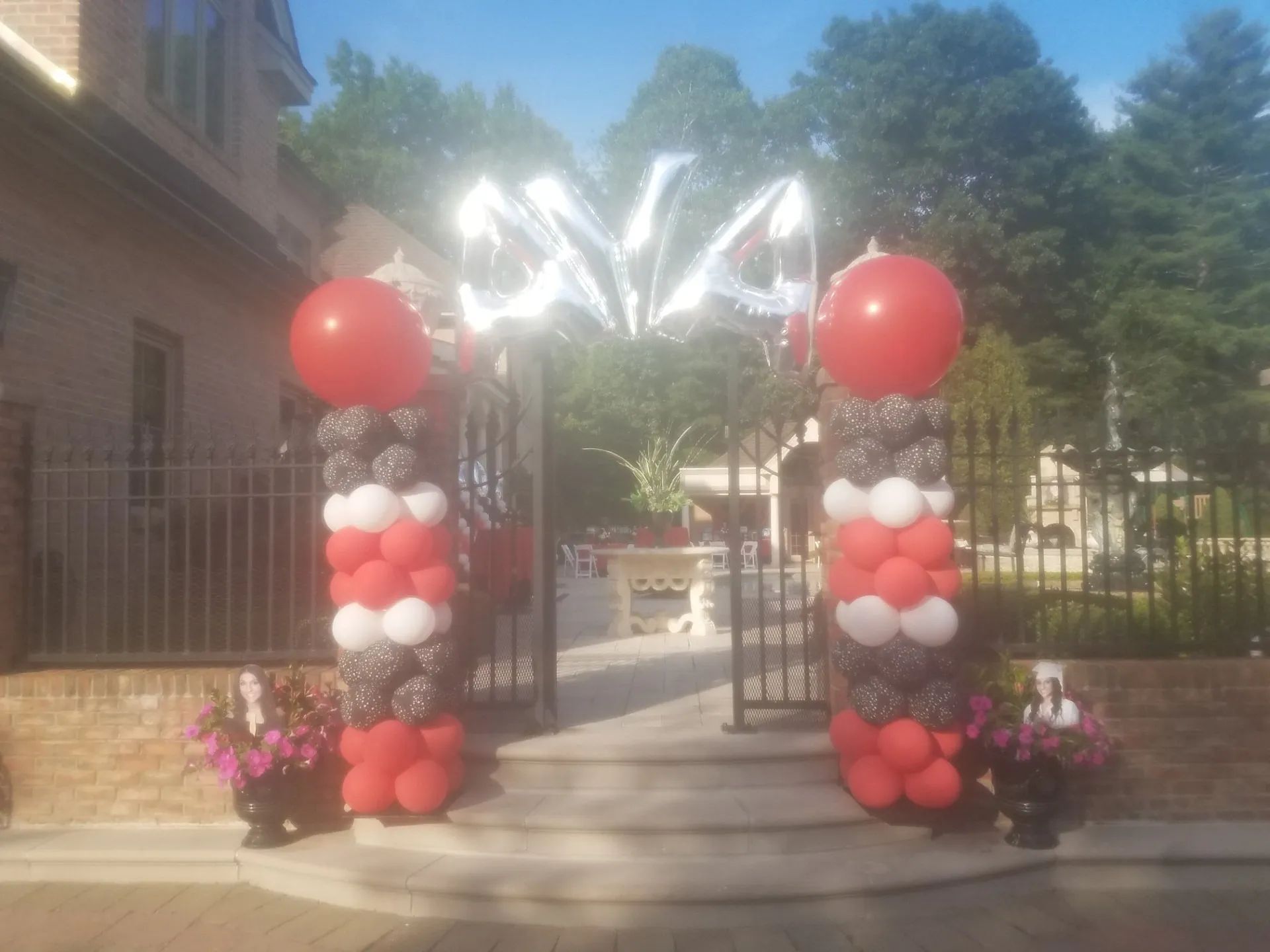 Balloon-decorated entrance gate with red and white columns, leading to a walkway and trees beyond