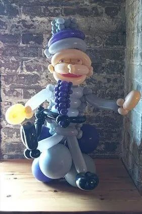 Balloon figure in a blue cap and white outfit, seated against a stone wall on a wooden floor.