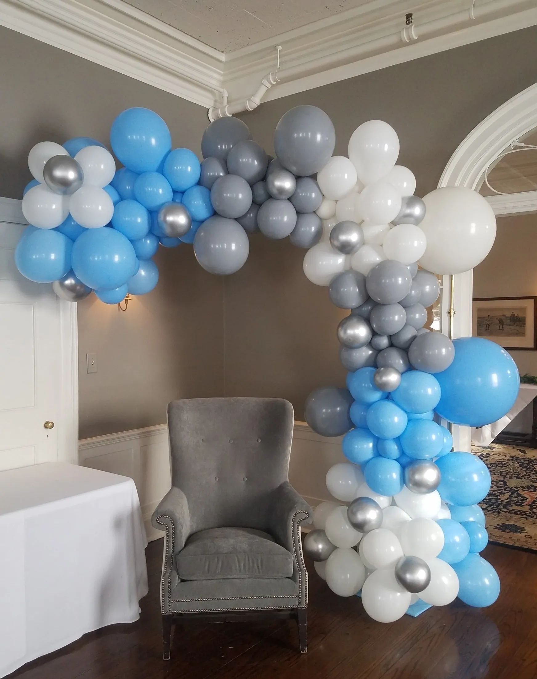 Blue, gray, and white balloon arch framing a gray armchair in a decorated indoor room.