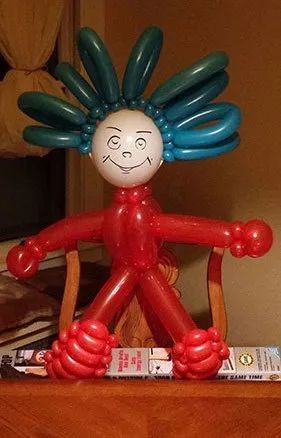 Balloon sculpture with blue hair, red limbs, and a painted face sitting on a chair