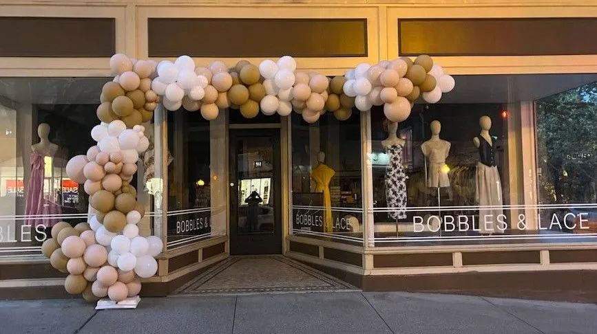 Balloon arch framing a storefront entrance with white, gold, and black balloons.