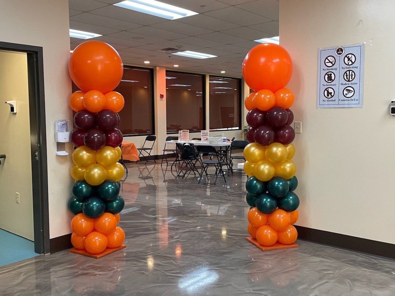Balloon columns framing a doorway in a hallway, with orange, maroon, gold, teal, and orange balloons.