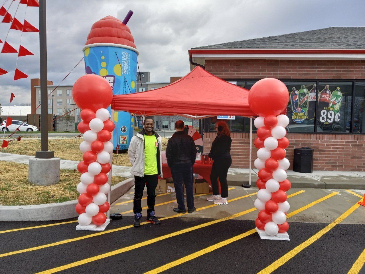 People under a red canopy at a store entrance, with red-and-white balloon columns and a cloudy parking lot.
