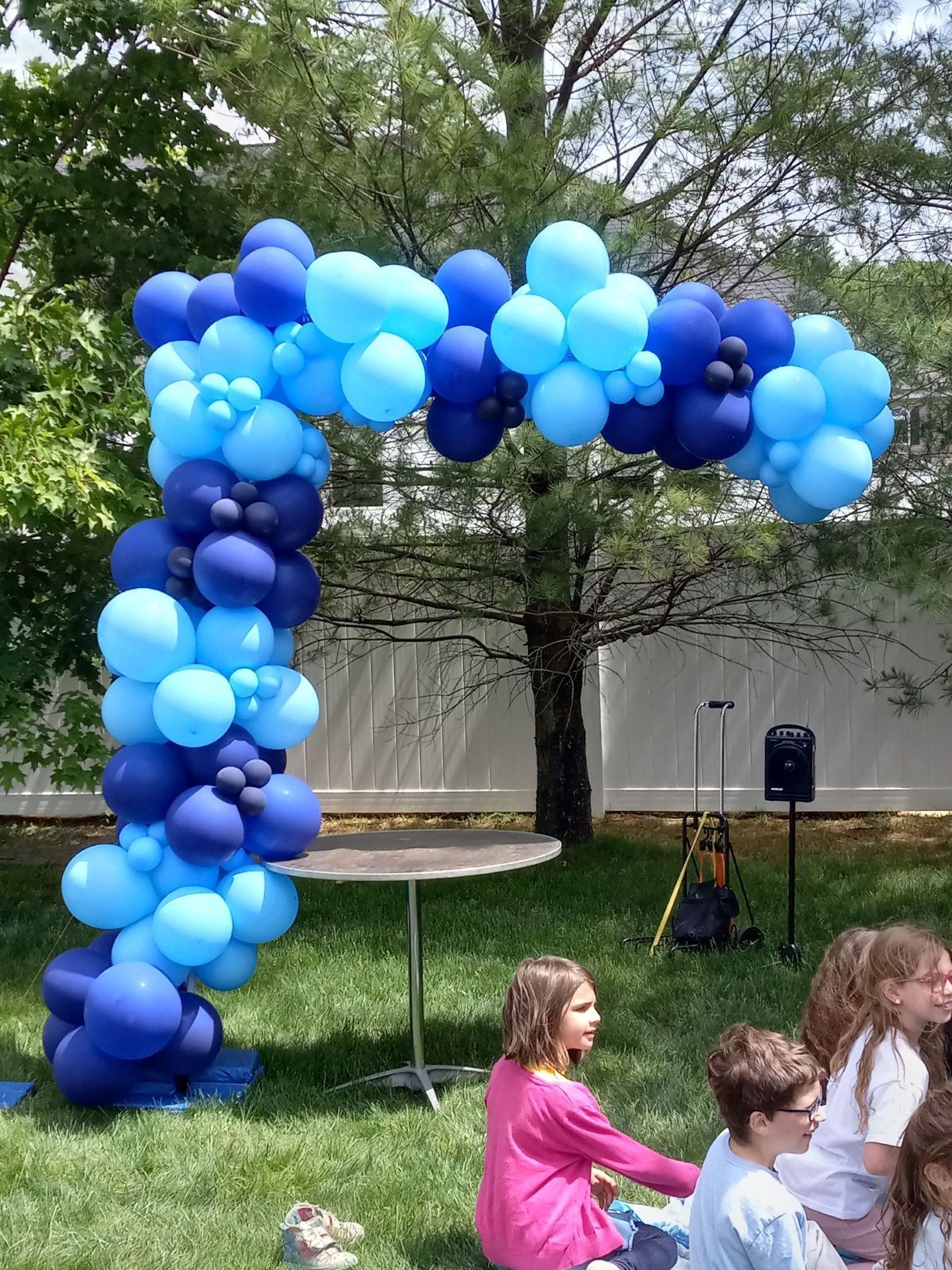 Blue balloon arch outdoors at a lawn gathering, with children in the foreground