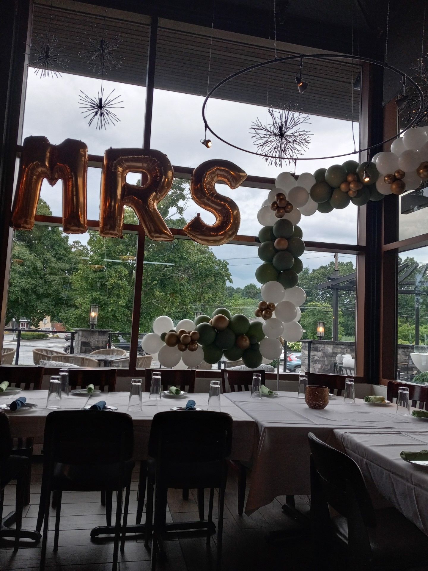 Decorated event table with gold “MRS” balloons and a white-and-green balloon arch by large windows