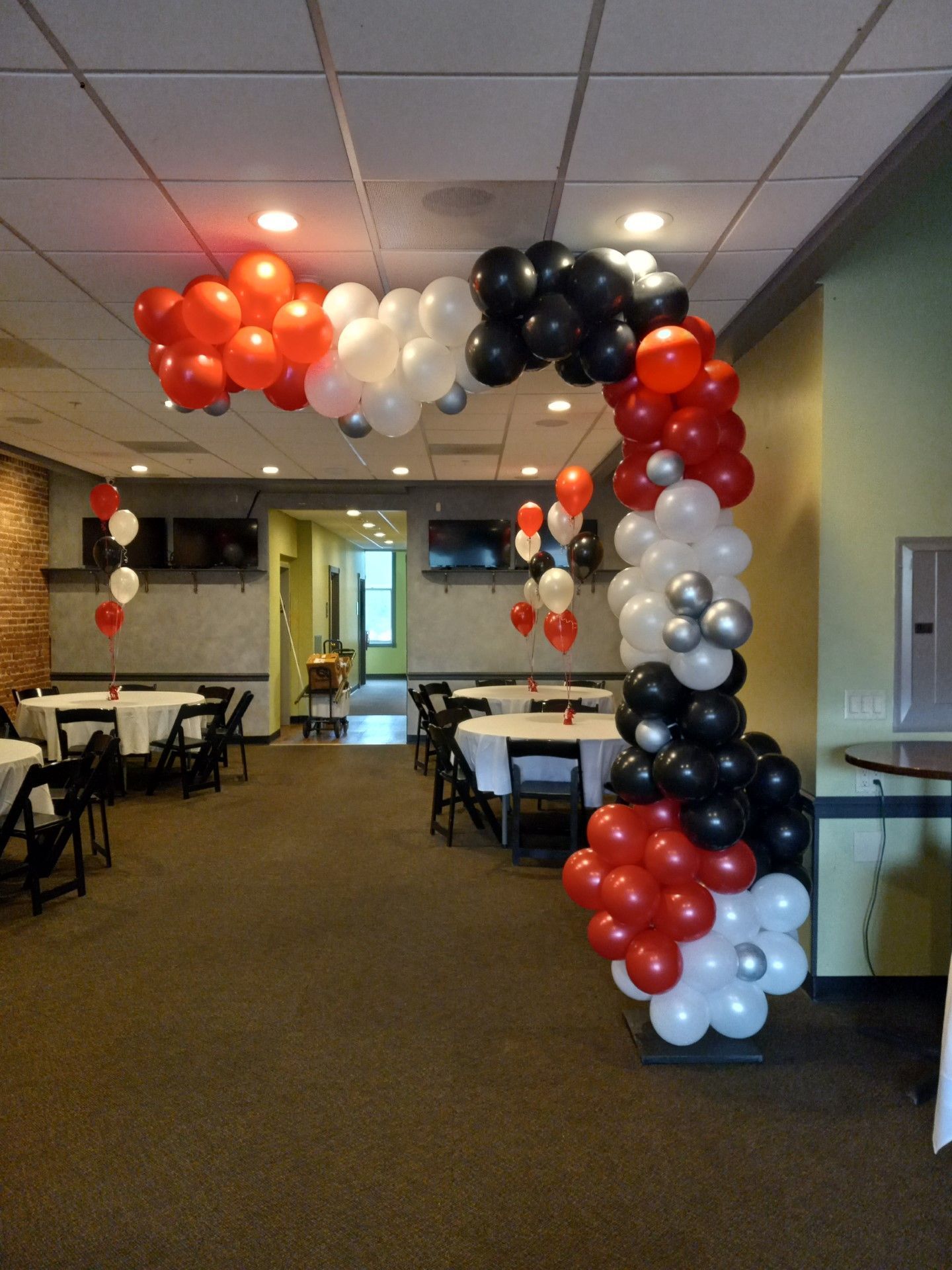 Red, white, and black balloon arch decorating a hallway event space with tables and chairs