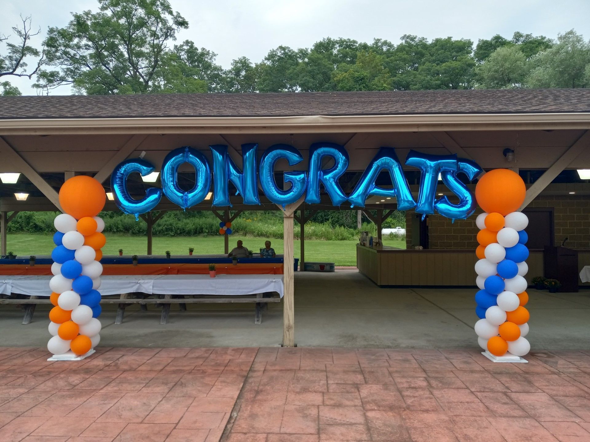 Blue “CONGRATS” balloon banner between orange, white, and blue balloon columns in an outdoor pavilion.
