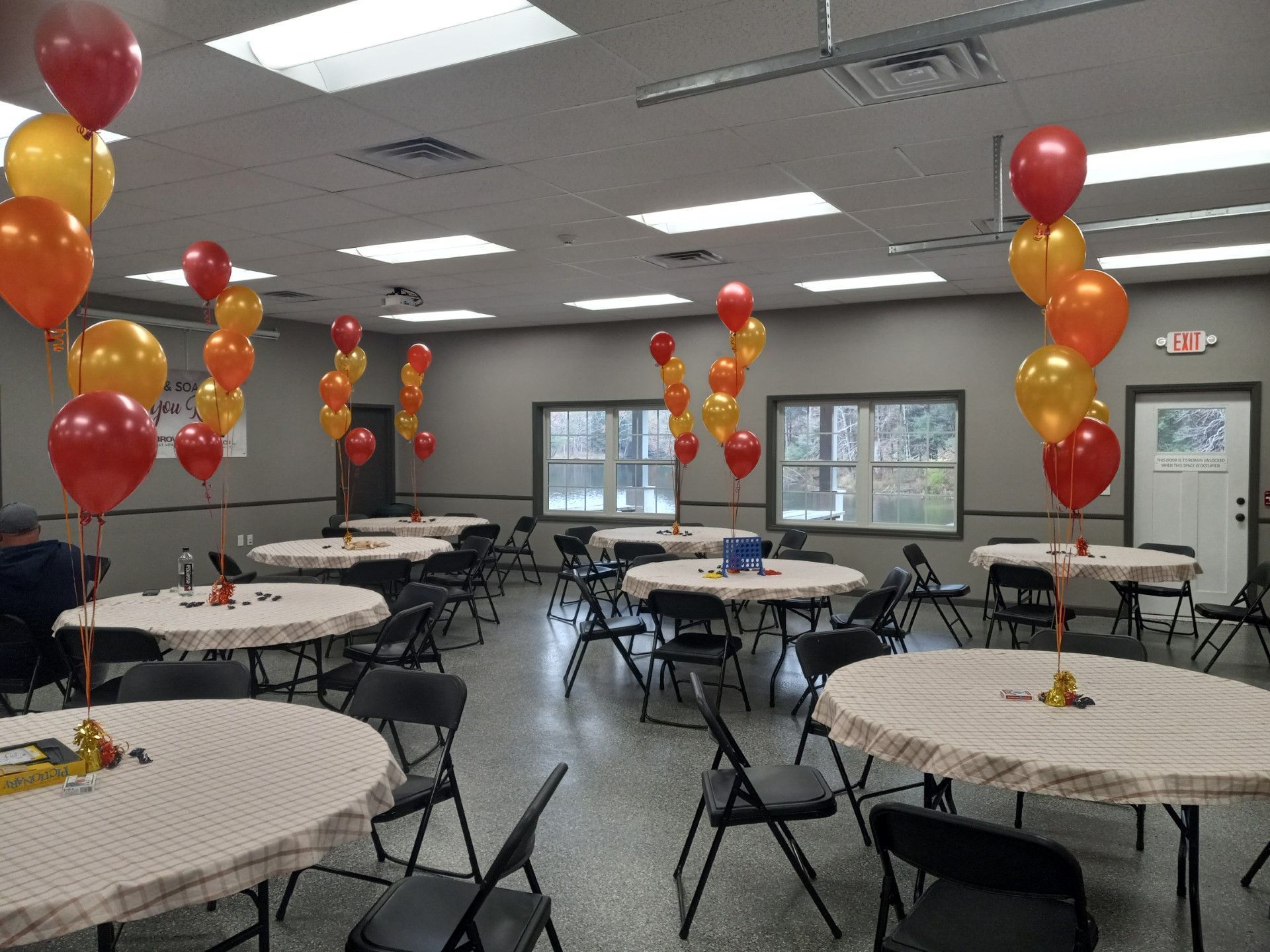 Balloon-decorated banquet hall with round tables and black folding chairs under fluorescent lights