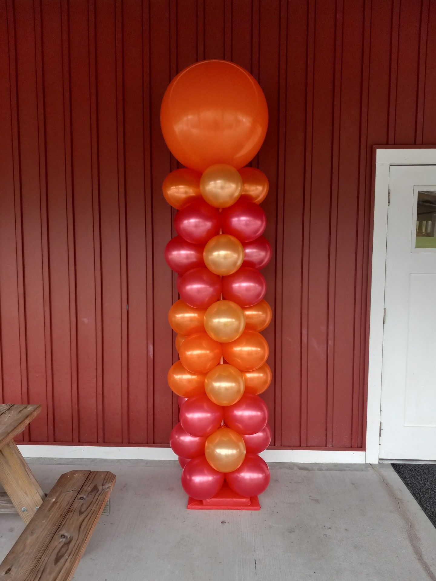 Tall balloon column in orange, gold, and red on a porch beside a red wall and white door.
