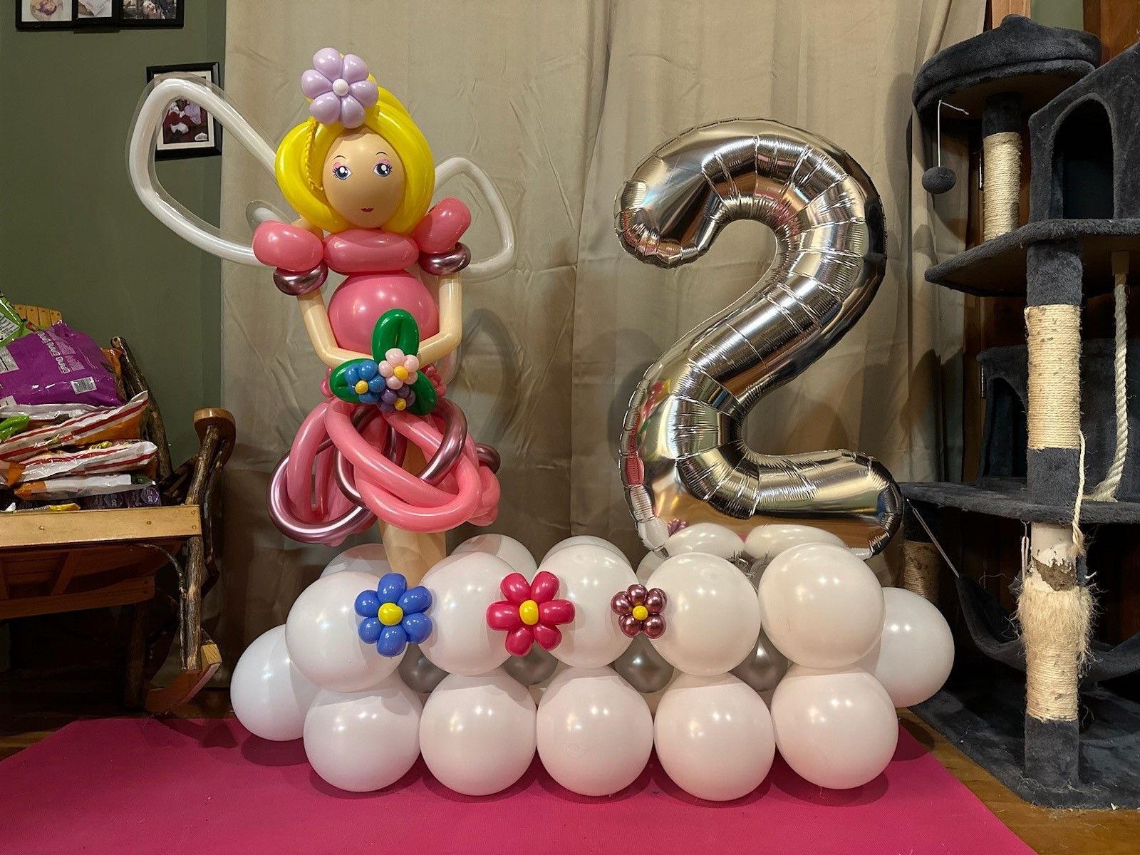 Balloon sculpture of a clown with a large silver “2” on a white balloon base, with flowers and a cat tree nearby