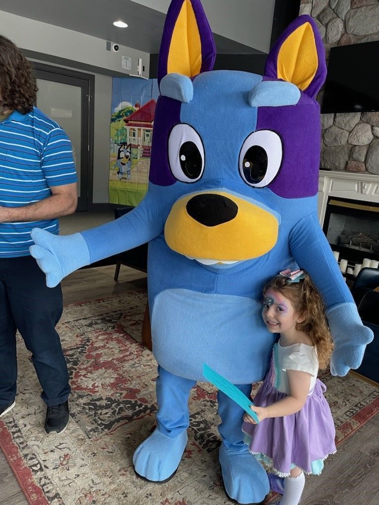 Blue dog mascot posing with a child in a purple dress inside a building lobby
