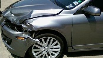 A silver car with a damaged front end is parked on the side of the road.