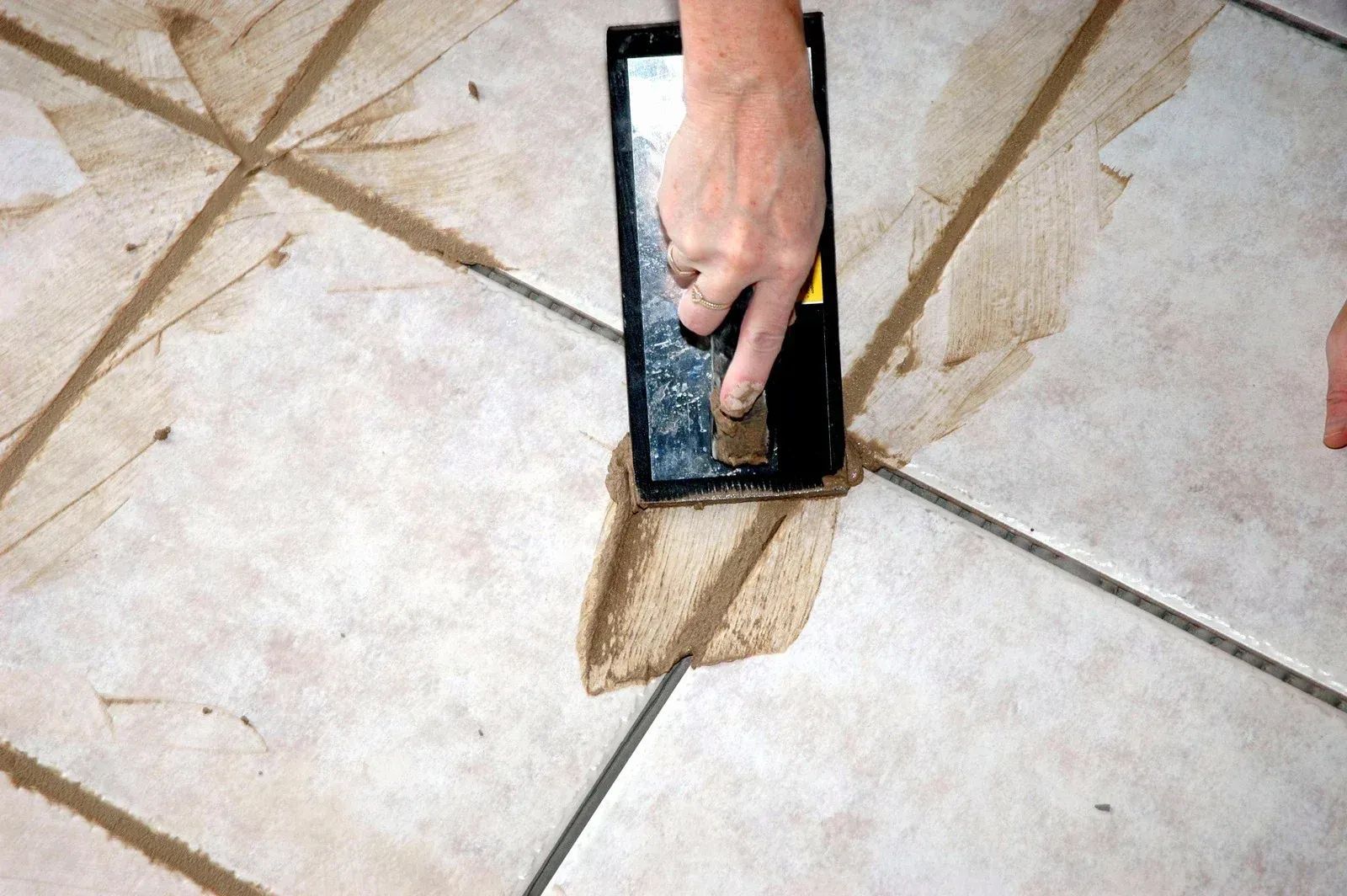 Person smoothing grout between beige floor tiles with a trowel