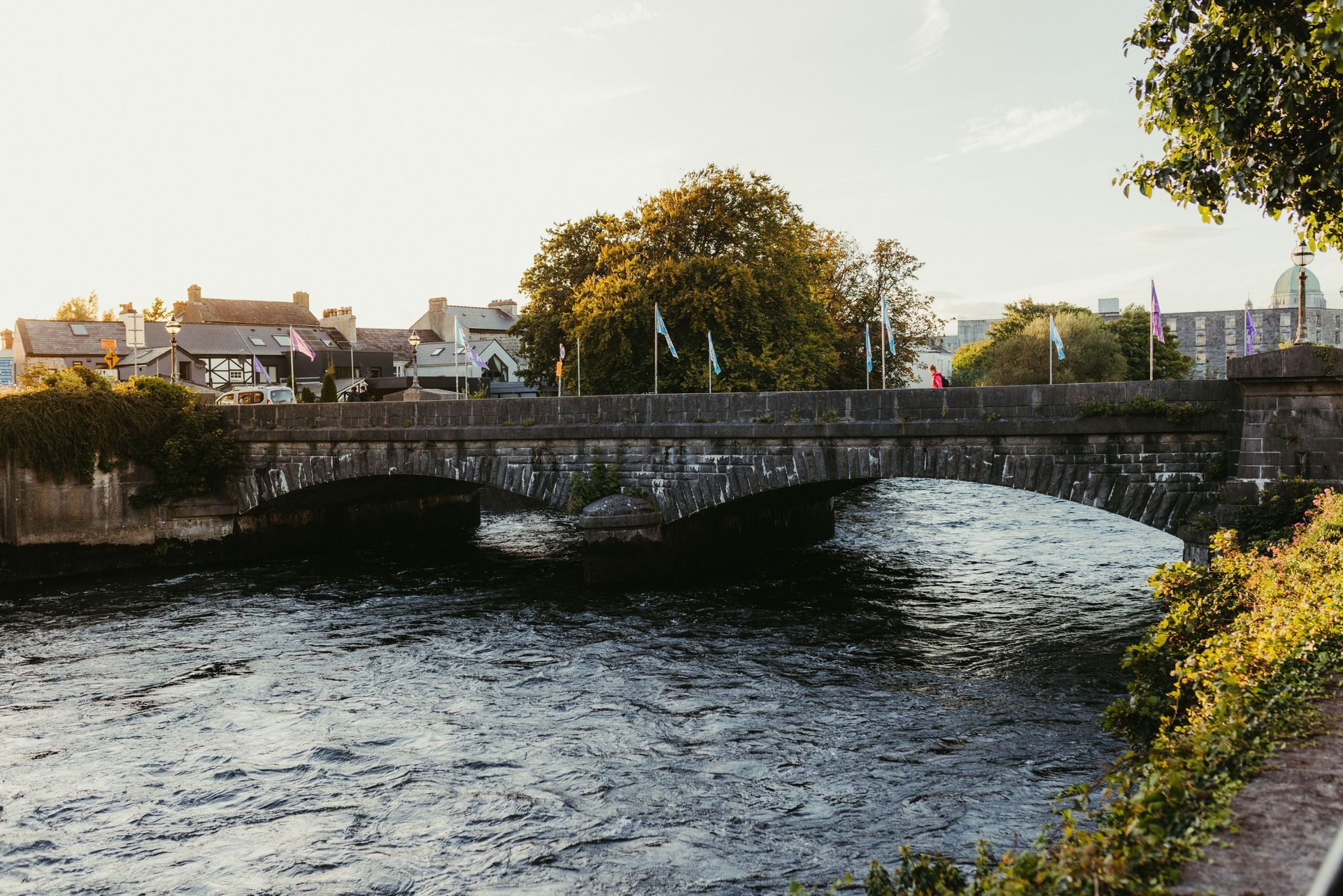 Stone bridge over a river at sunset, with houses and trees along the banks.