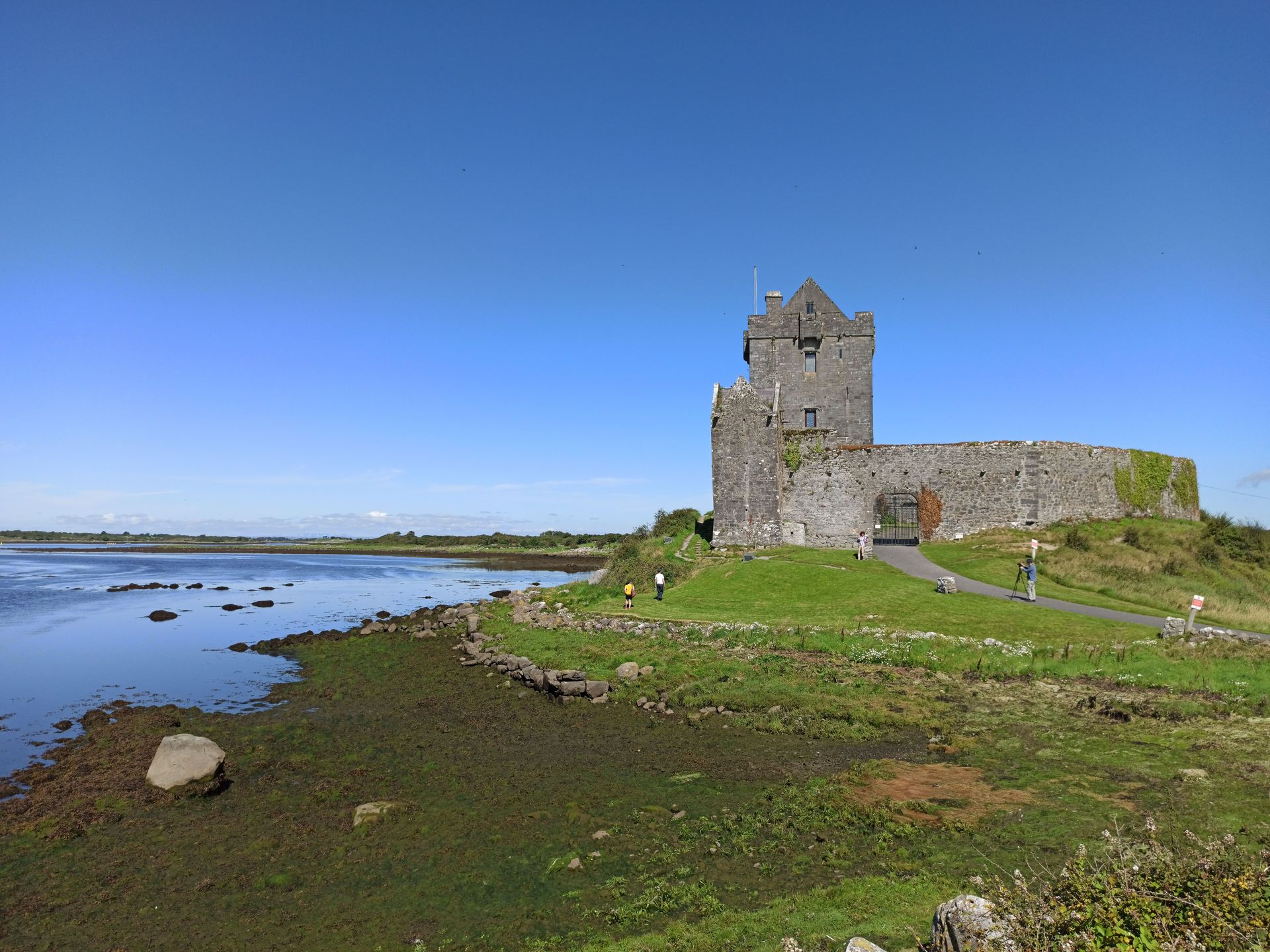 Stone castle ruin on a grassy shoreline beside calm water under a clear blue sky.