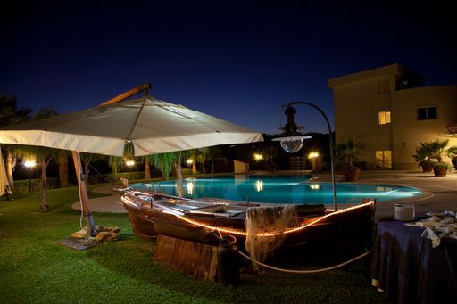 Scena notturna a bordo piscina con un bar a forma di barca, un ombrellone illuminato e un edificio.