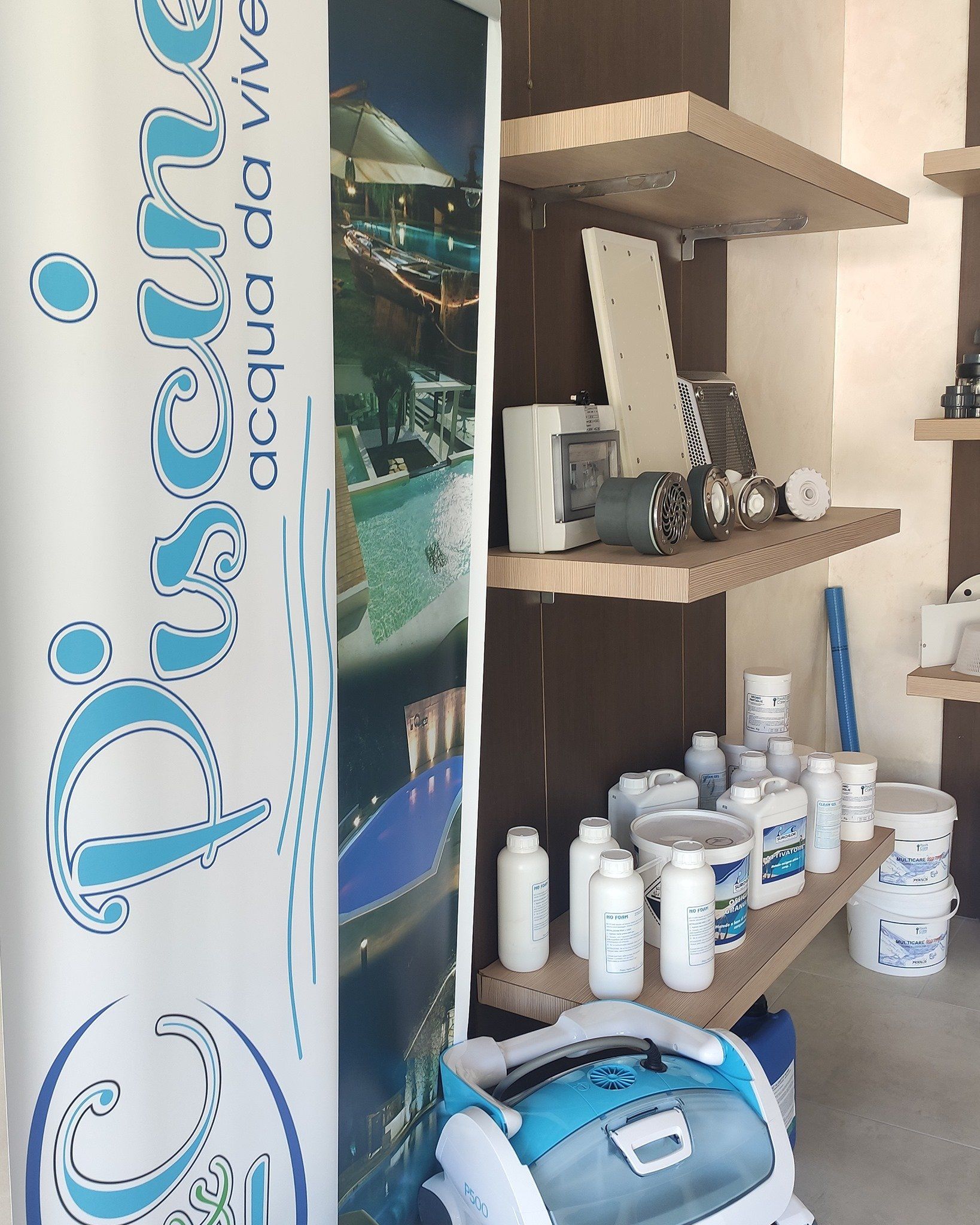Forniture per piscine sugli scaffali, tra cui prodotti chimici e un pulitore robotizzato, vicino a uno striscione.