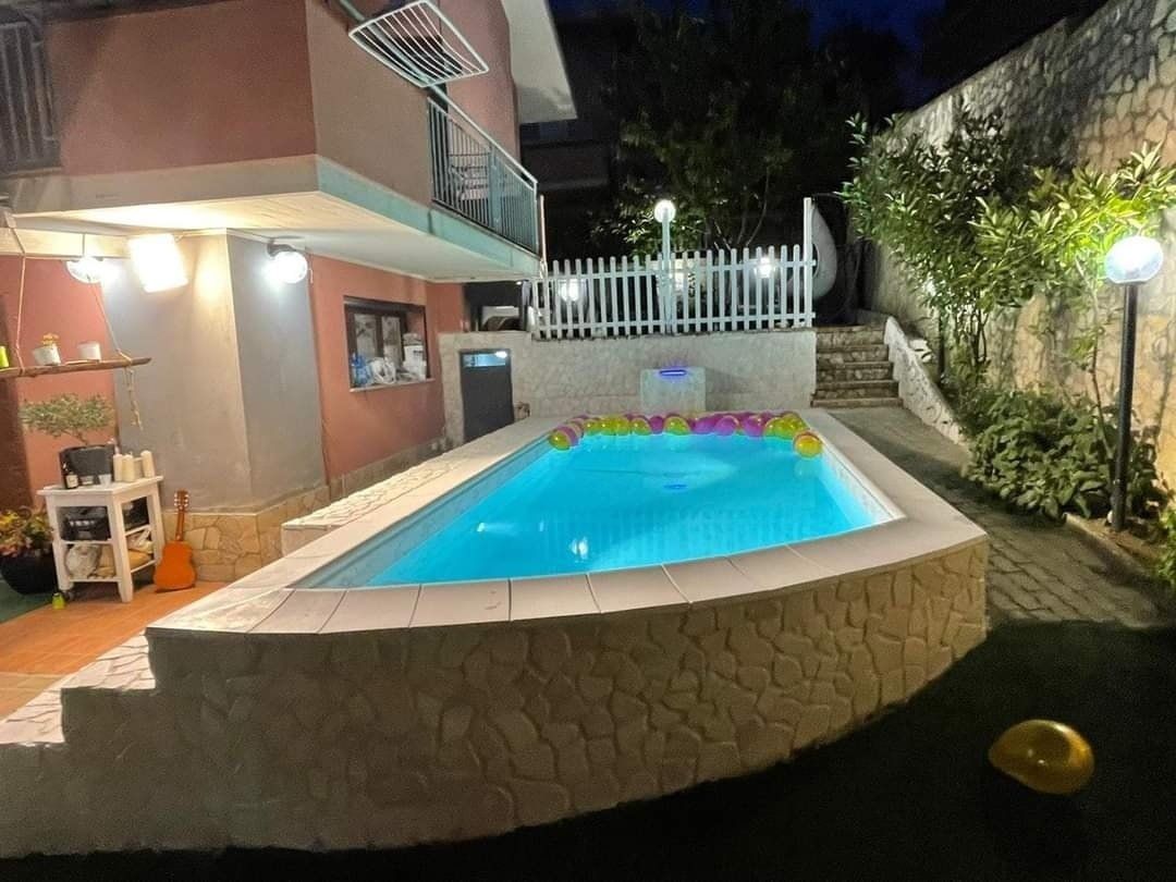 Piscina davanti a una casa, illuminata di notte. Una scalinata conduce a una staccionata bianca. Erba verde.