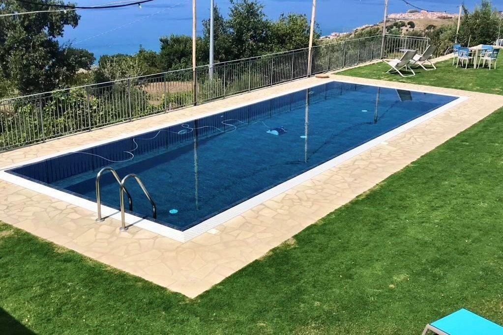 Piscina esterna rettangolare con scaletta, circondata da prato verde e recinzione, con vista sul mare.