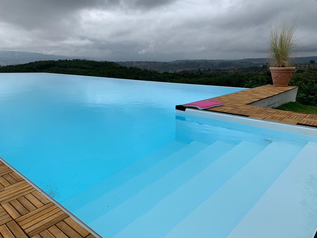 Piscina a sfioro con vista sulle colline sotto un cielo nuvoloso. Terrazza in legno, piante in vaso e acqua azzurra.