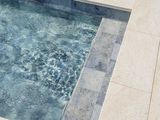 Primo piano di una piscina con acqua increspata, bordo in pietra grigia e rivestimento in piastrelle color marrone chiaro.